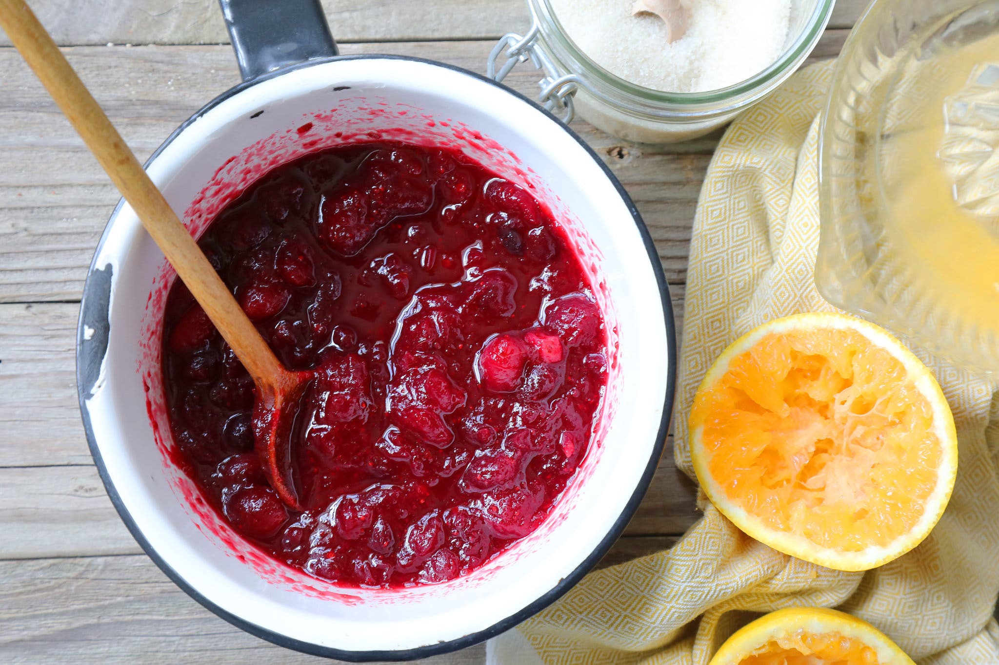 An enameled pot holds just-cooked cranberry sauce. To the right are halved, freshly squeezed oranges, and above is a bowl with sugar. 