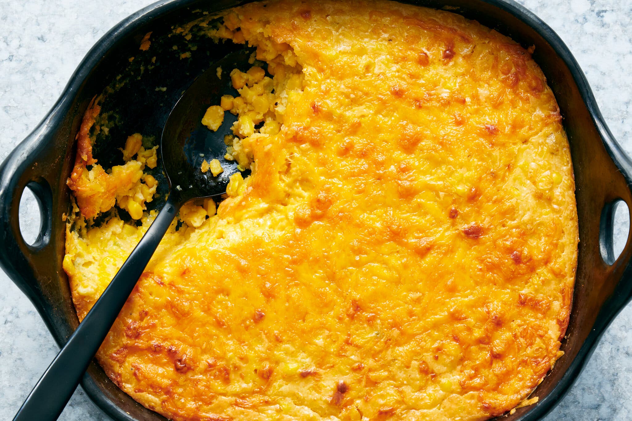 A black skillet holds a dark yellow corn casserole with cheese melted on top.