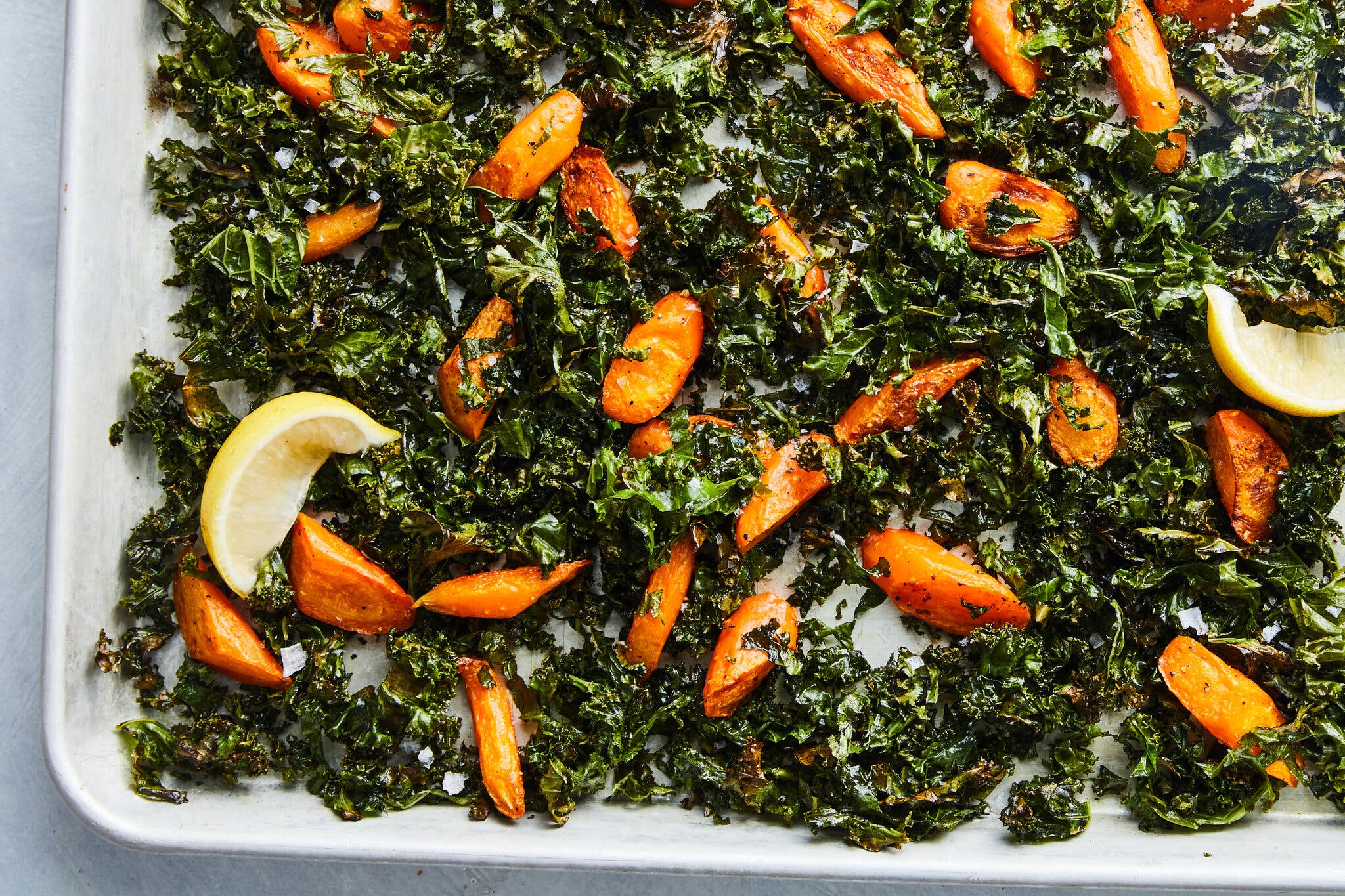 A sheet pan is covered with kale and carrot slices, plus a lemon wedge.