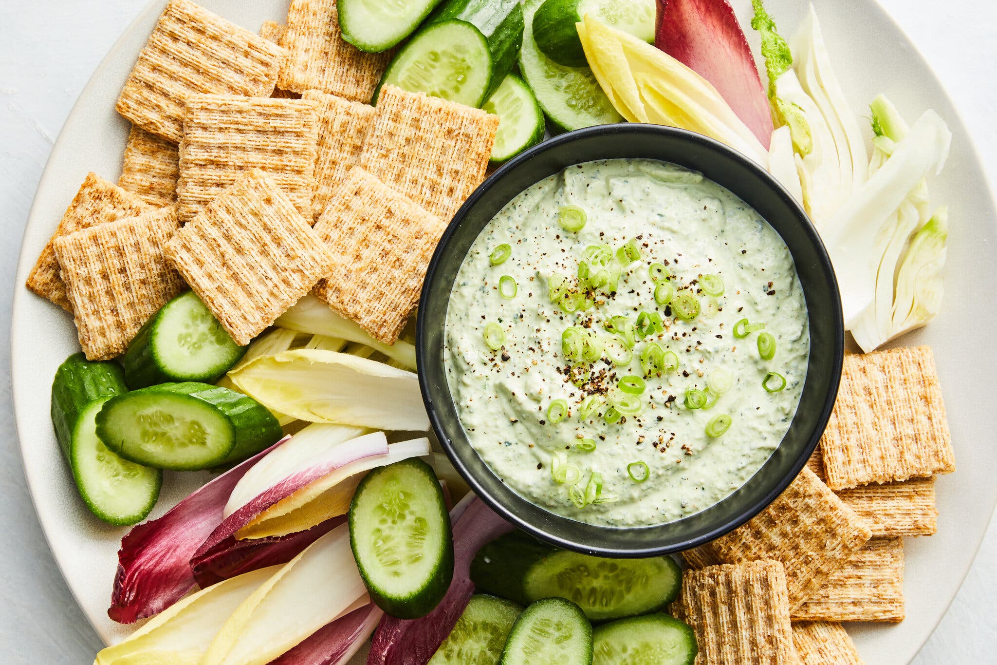 A green dip topped with scallions is surrounded by sliced vegetables and wheat crackers.