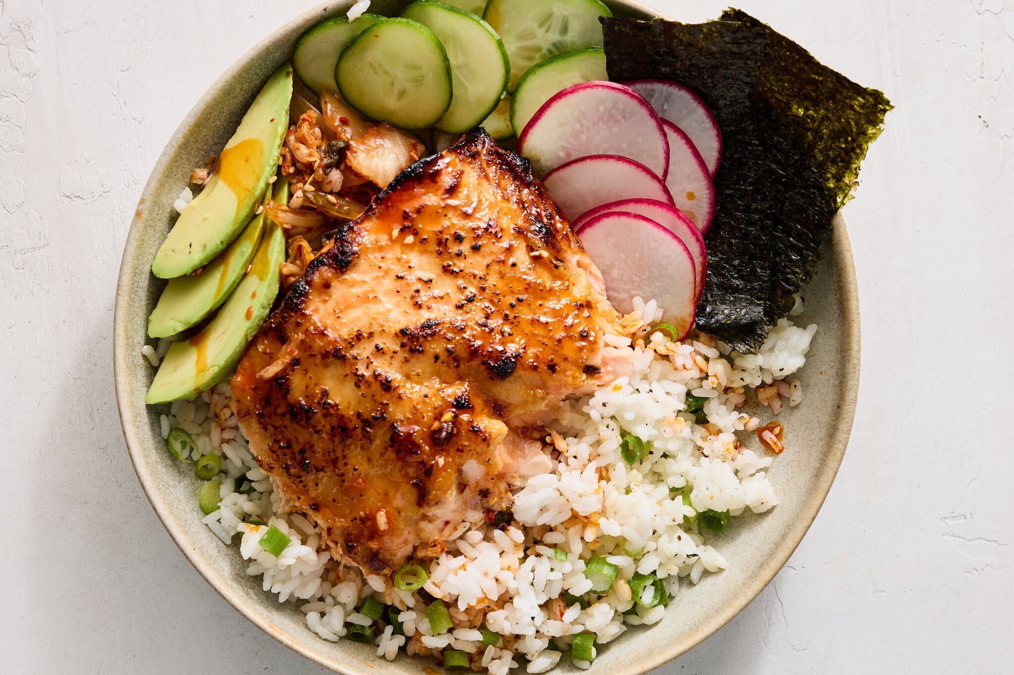 An overhead image of rice and salmon in a bowl, with sliced avocado, cucumbers and radishes.