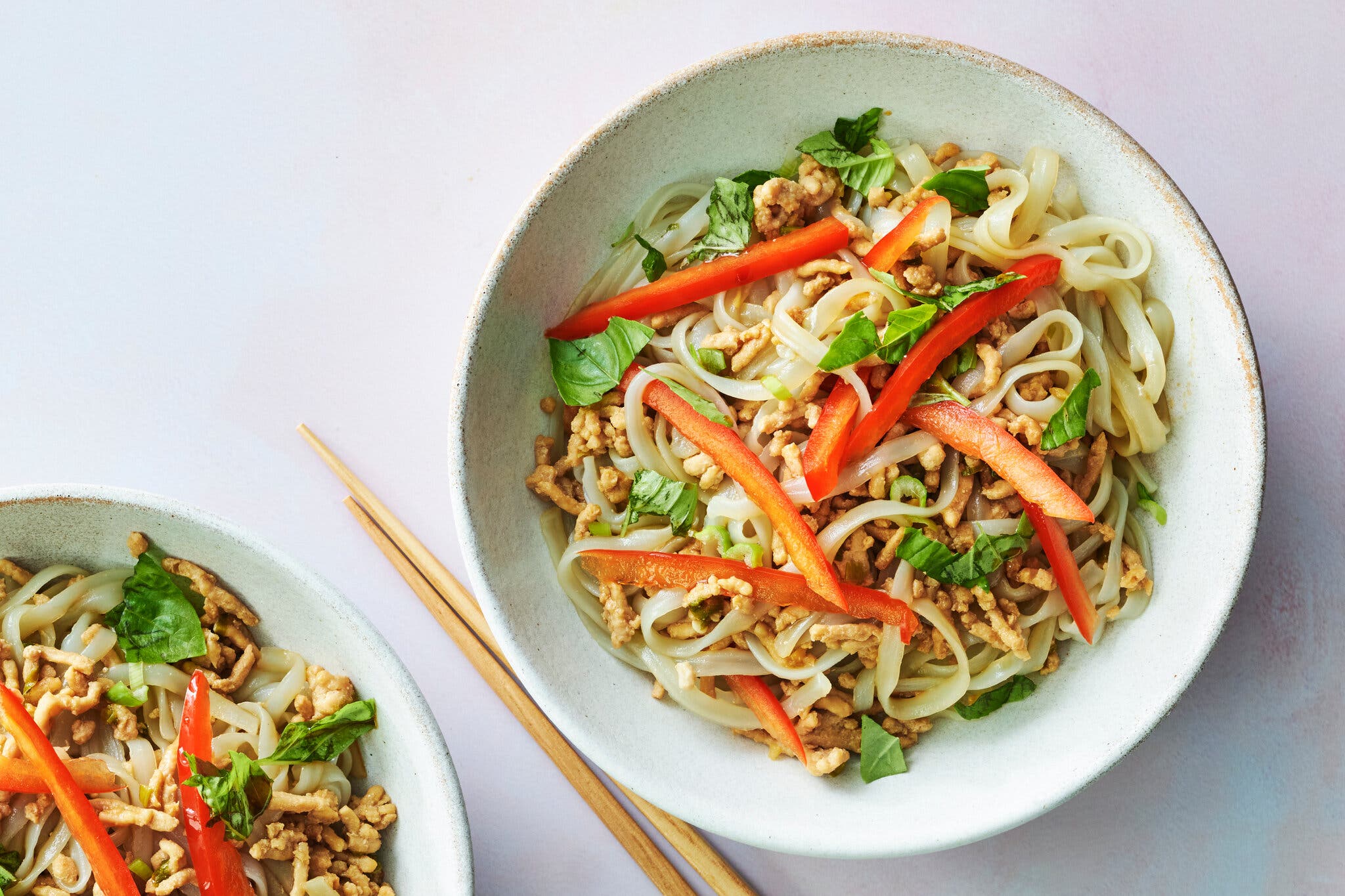 Two white bowls are filled with noodles topped with ground chicken, herbs and sliced red peppers.