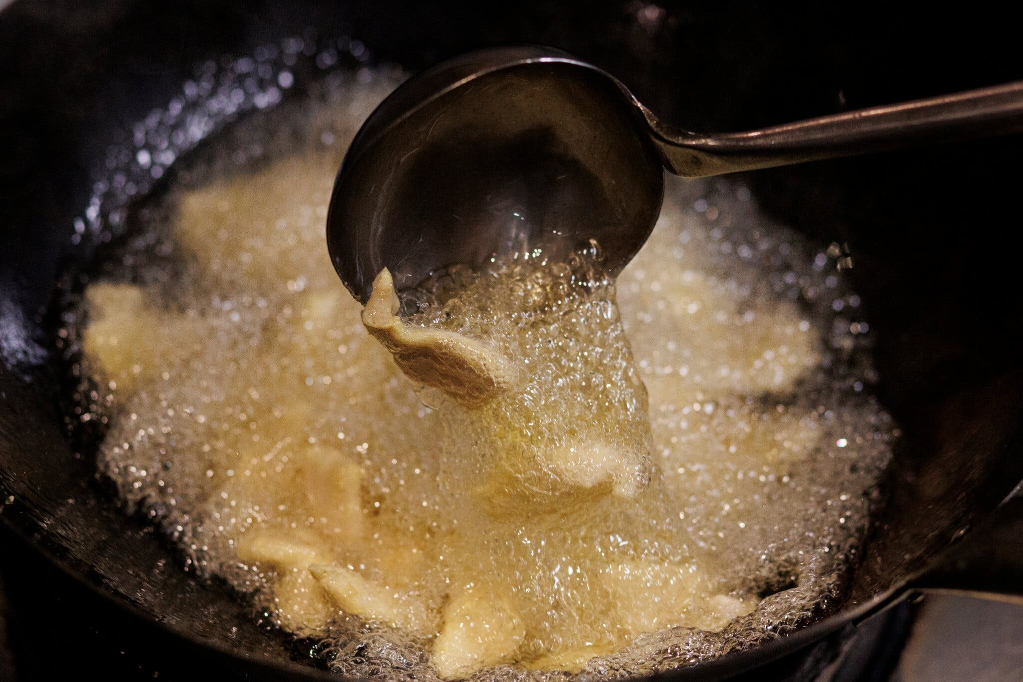 Oil bubbles in a black wok filled with chicken slices. A ladle is spooning out some of the meat.