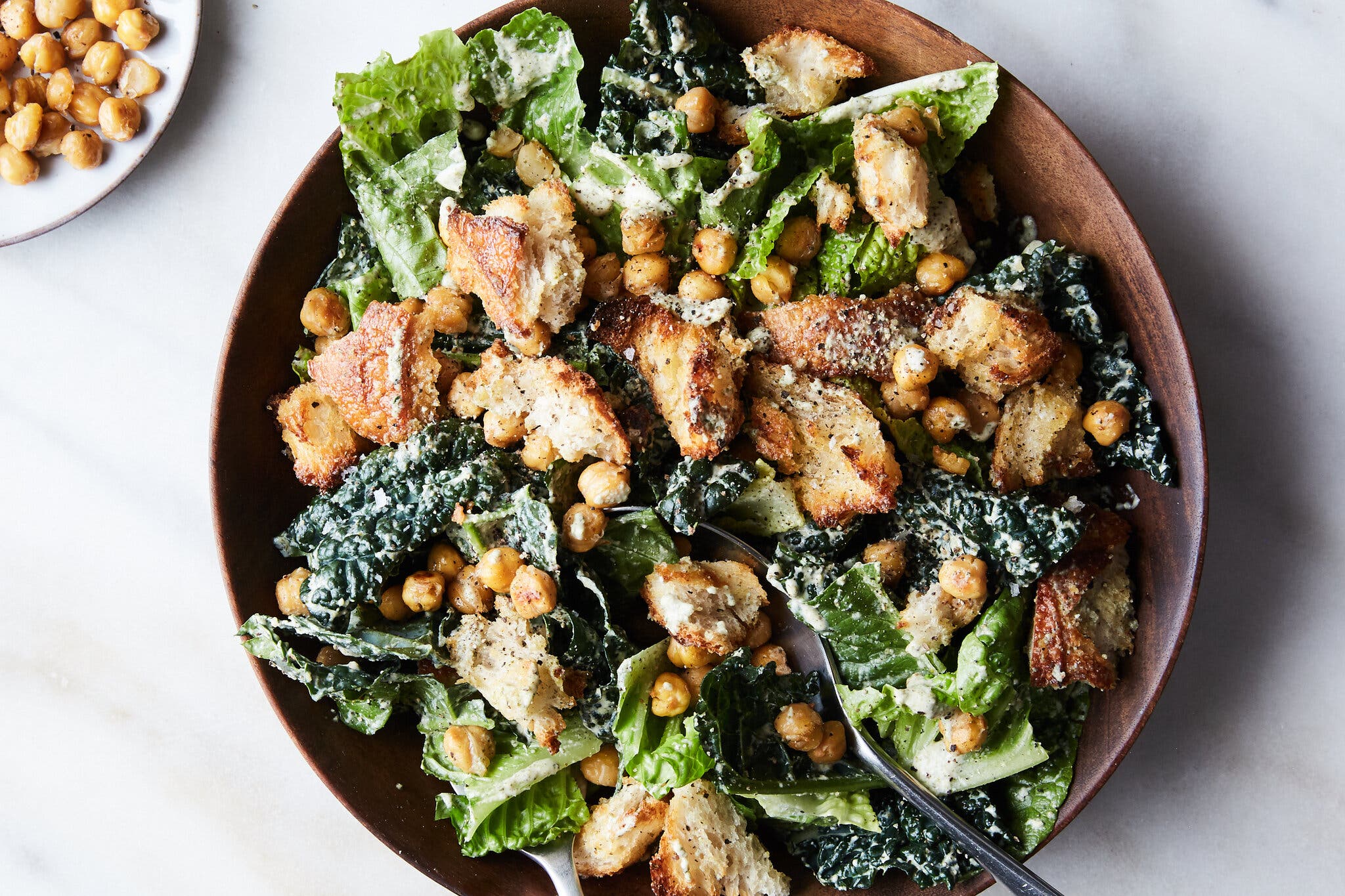 A wooden bowl of salad with romaine lettuce, kale, chickpeas and hand-torn croutons, along with a large serving spoon.