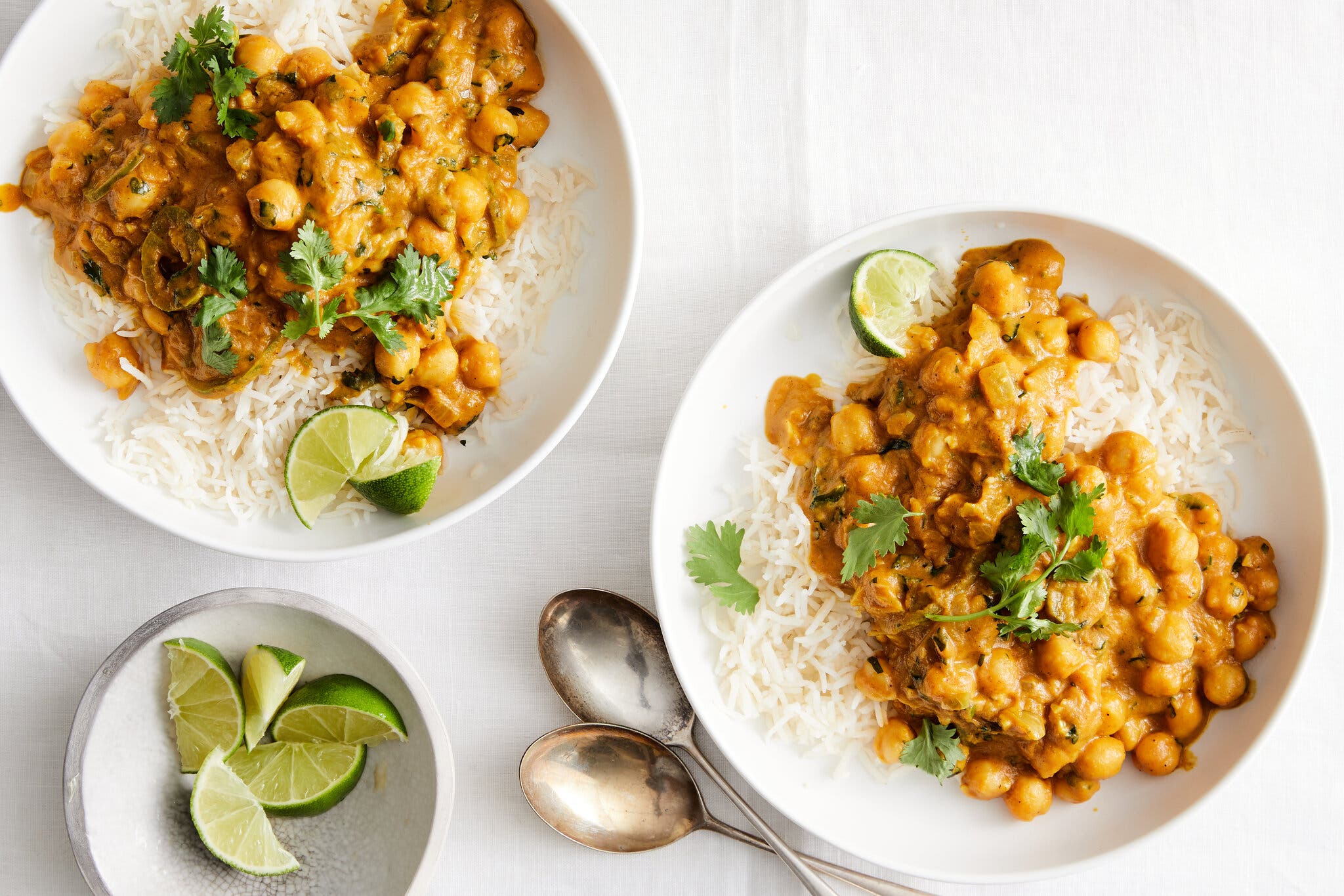 Two bowls of white rice, topped with chickpea curry and fresh cilantro, along with a small bowl of lime wedges.