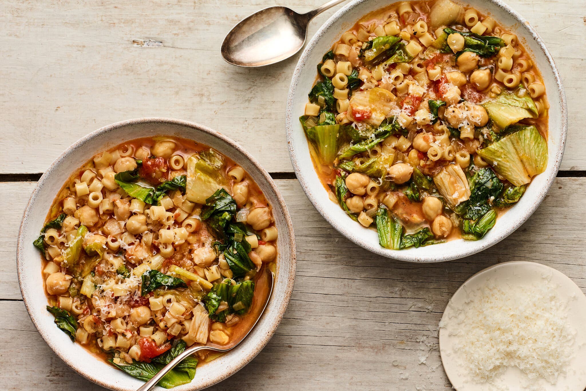 Two bowls of stew, on a wooden surface, with chickpeas, escarole, tomatoes and grated pecorino. 