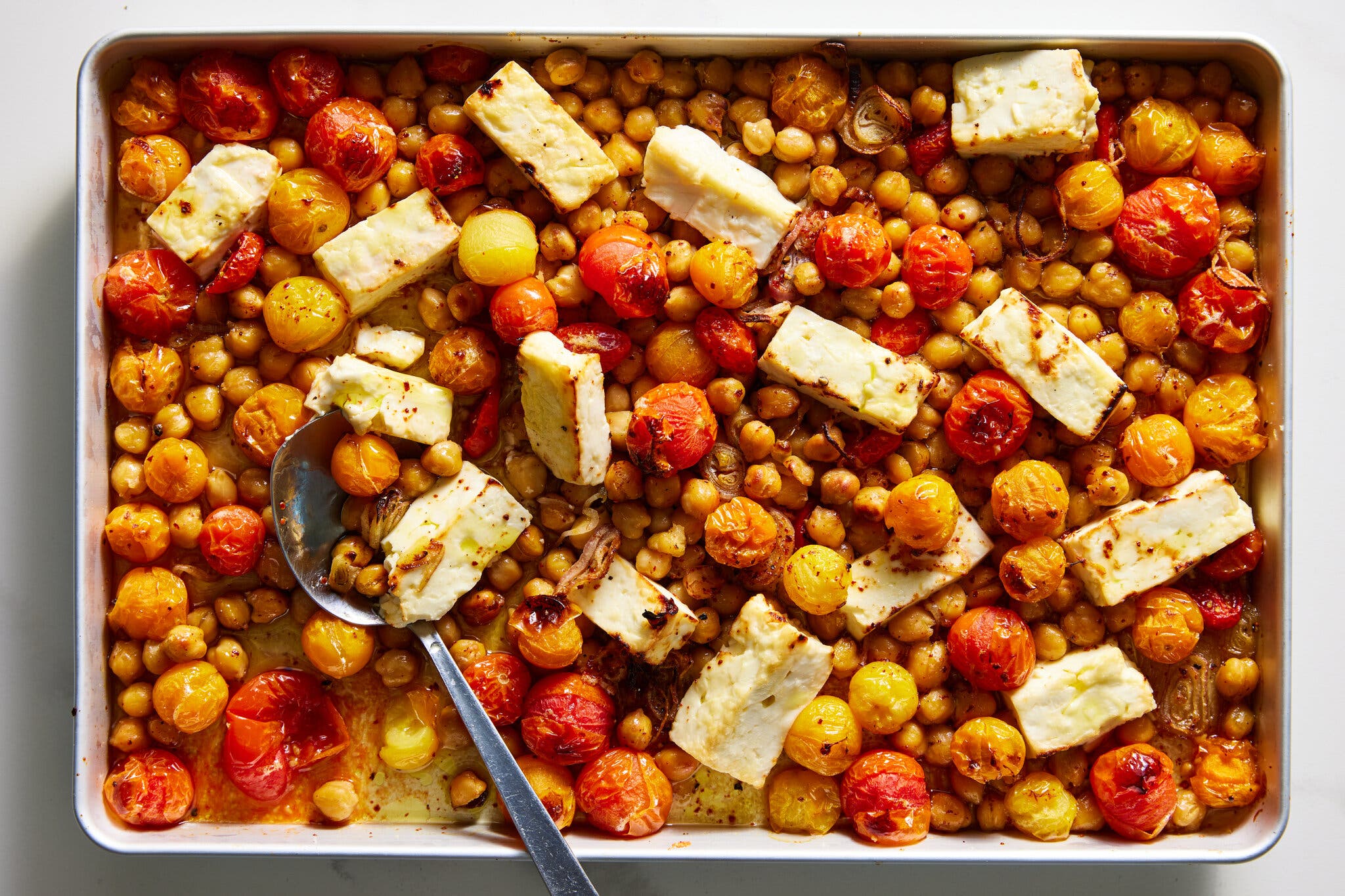 A sheet pan with chickpeas, cherry tomatoes and thick slices of feta, along with a serving spoon.