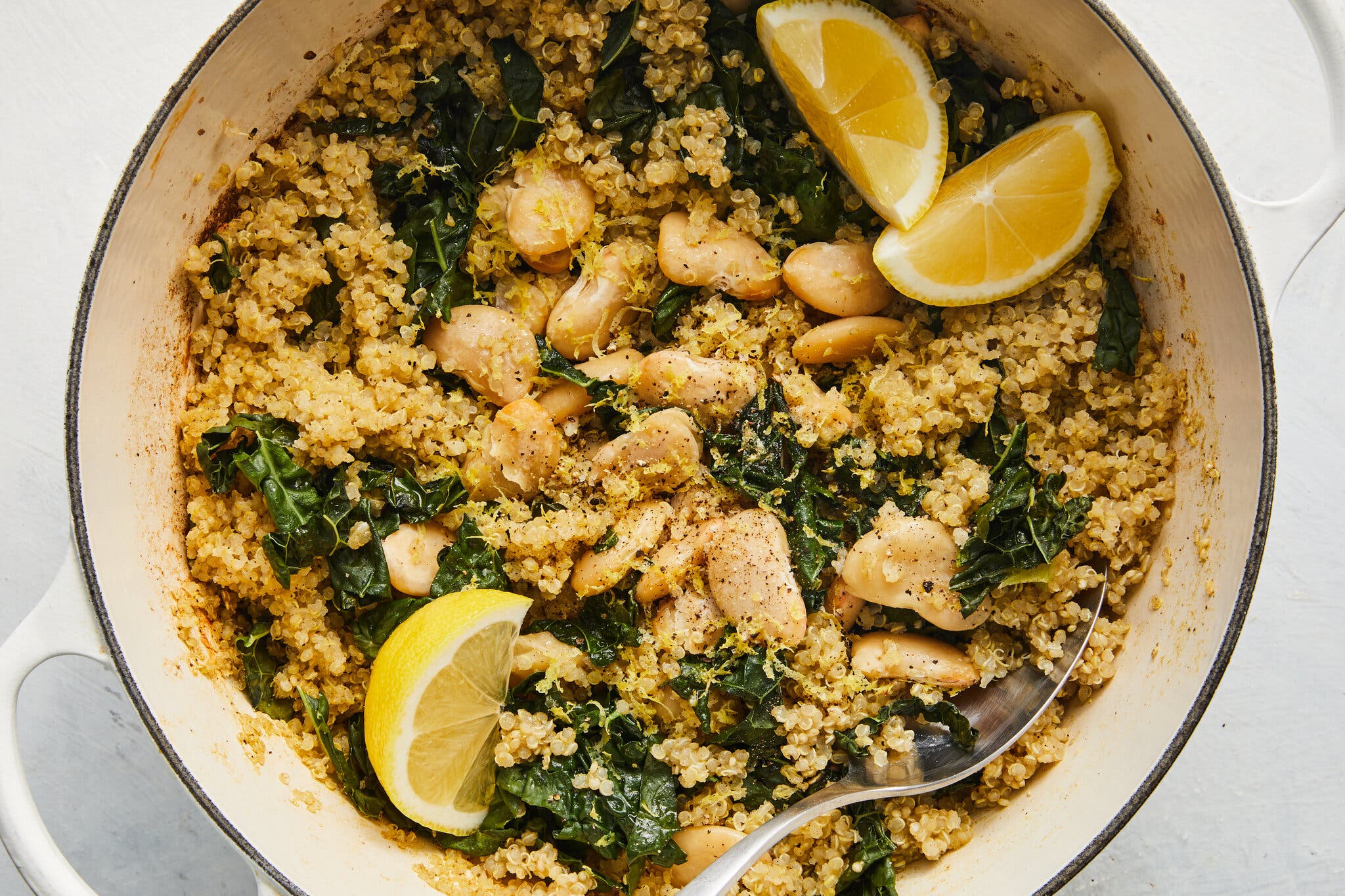 A large white Dutch oven is filled with quinoa, white beans, kale and lemon wedges.