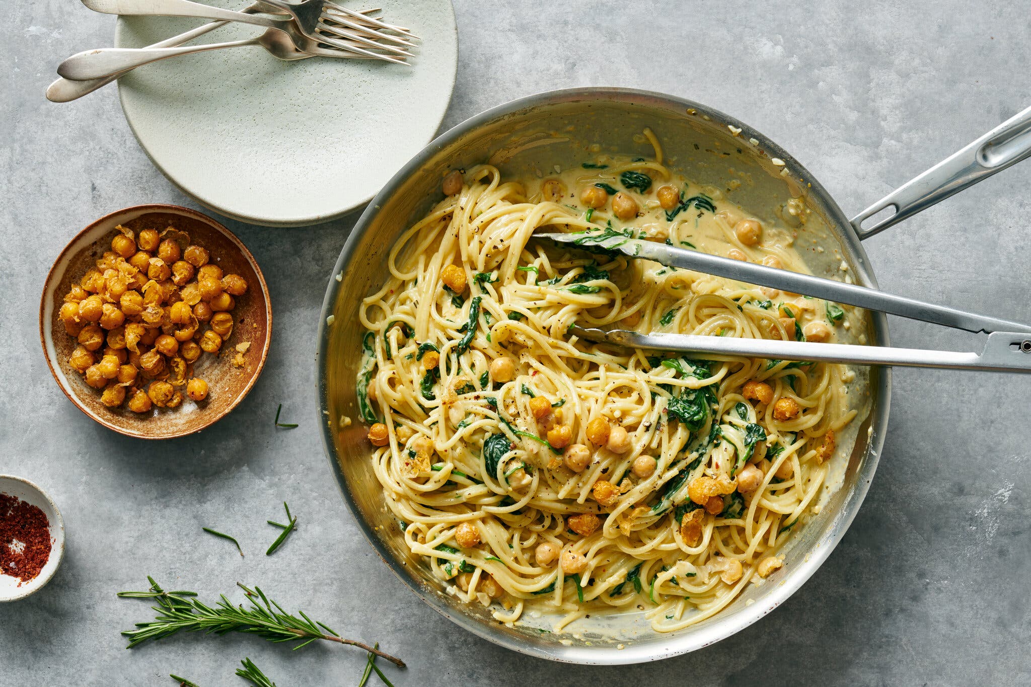 A from-above shot of spaghetti, in a creamy sauce with chickpeas and spinach, in a pan, along with a pair of tongs. To the left, a stack of plates with forks, a small bowl of spiced chickpeas and a sprig of rosemary.